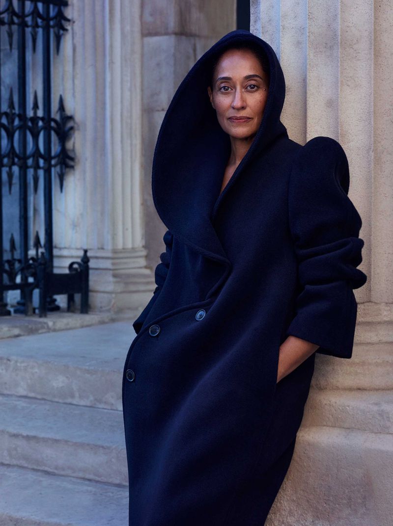 A woman with dark hair wearing a navy blue hooded coat leans against a marble pillar outdoors with a wrought iron fence in the background.