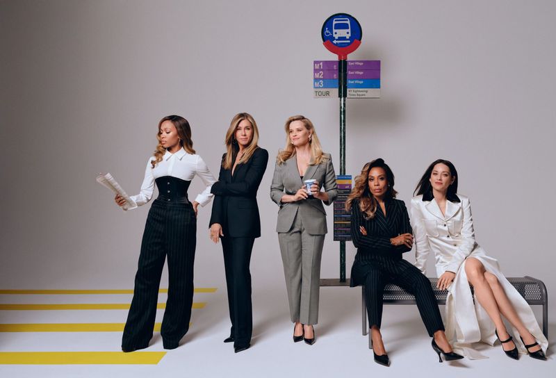 Five women in business suits pose at a stylized bus stop in a studio, showcasing fashion and professional style against a neutral background.