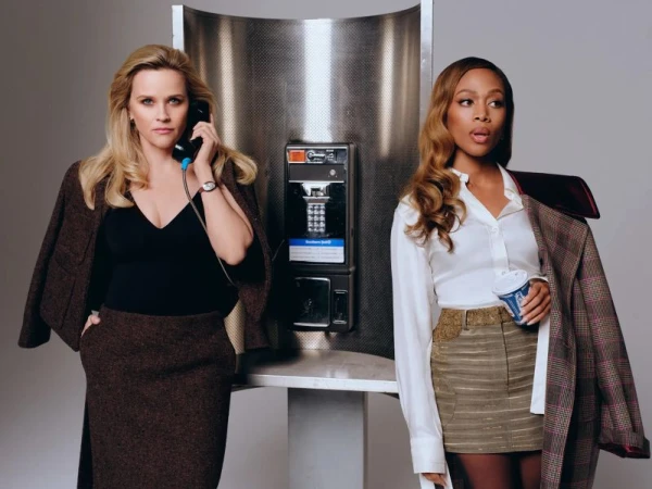 Two women in business attire stand by a vintage phone booth; one speaks on the receiver, the other holds a drink and a coat.