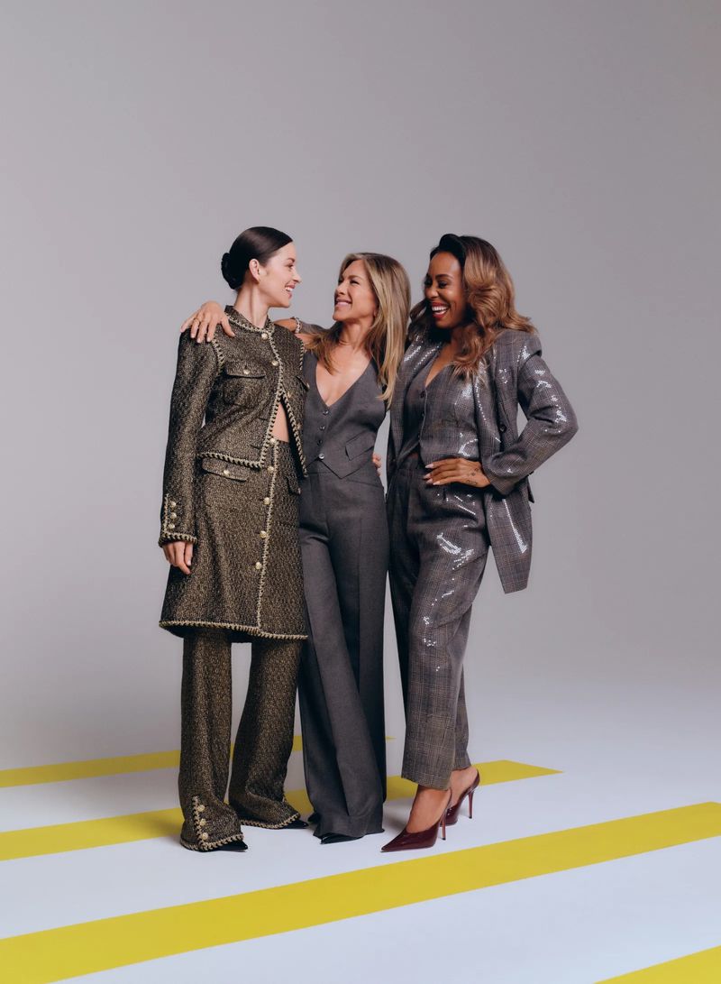 Three women stand together in suits, two embrace, on a yellow and white striped floor in a studio with grey backdrop, medium shot.