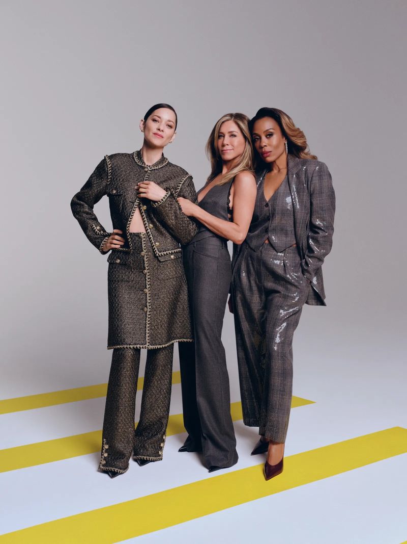 Three women stand together in studio wearing tailored suits in gray tones. One woman wears a sequined suit, another a classic suit.