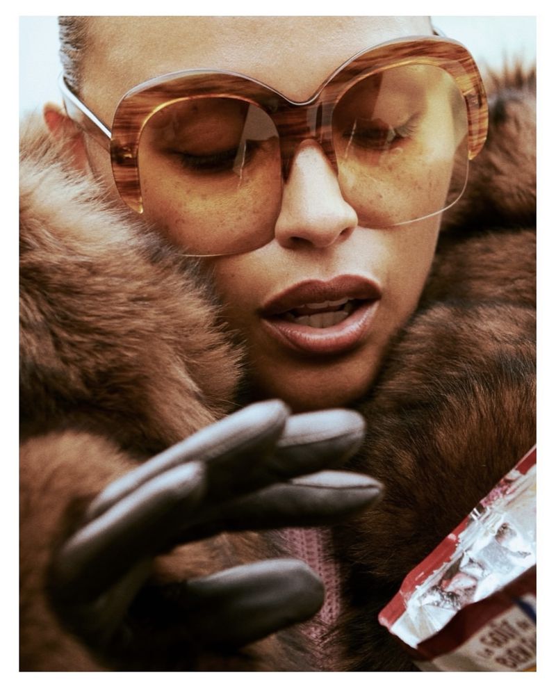 Close-up portrait of a woman in a fur coat and sunglasses holding a bag of chips, shot in a photo studio with controlled lighting.