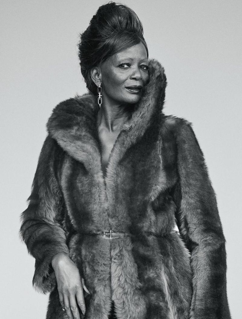 Elegant mature woman with a stylish updo and statement earrings posing in a luxurious fur coat against a neutral background.