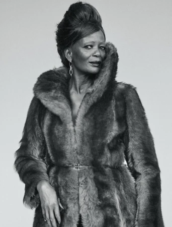 Elegant mature woman with a stylish updo and statement earrings posing in a luxurious fur coat against a neutral background.
