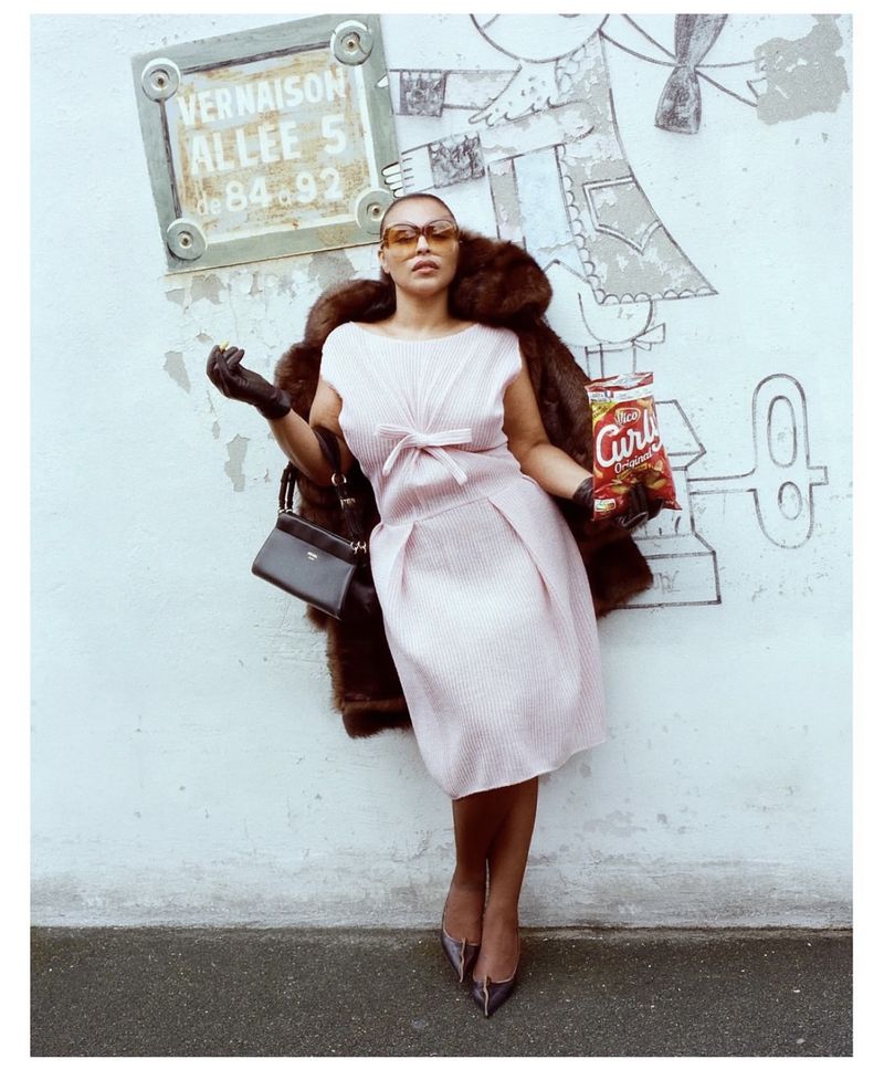 Woman in pink dress and fur coat poses next to graffiti wall with Curly snack, wearing sunglasses and black gloves in an urban setting.