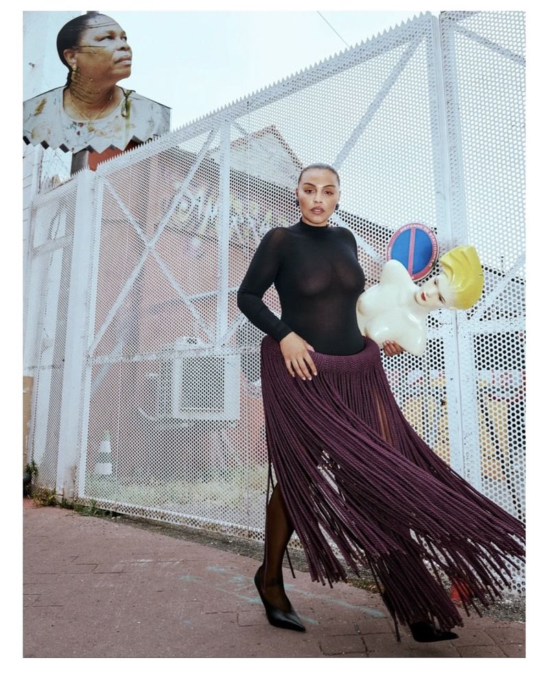 A woman poses with a mannequin head in front of a gated fence, wearing a sheer black top and a purple skirt with tassels.