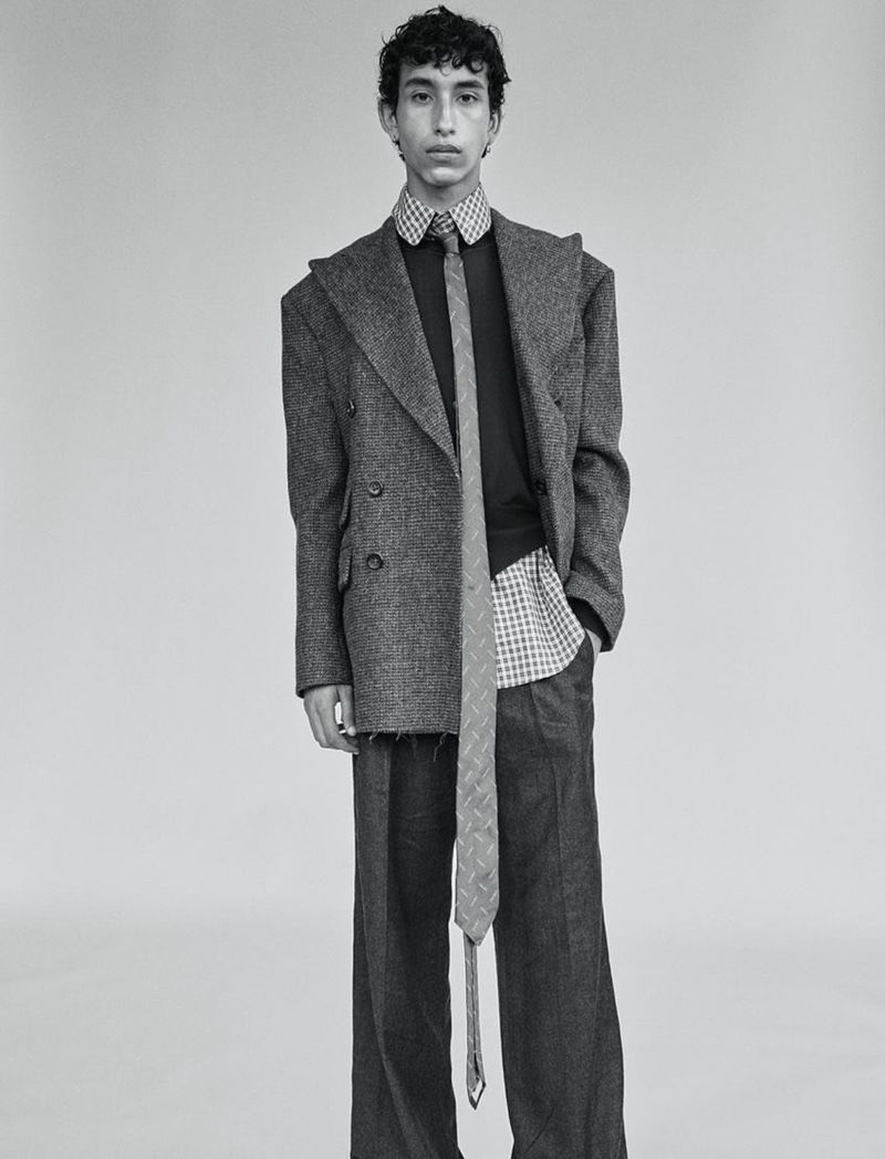 Monochrome studio shot of a young adult posing in an oversized grey suit with a patterned shirt and tie, set against a neutral backdrop.
