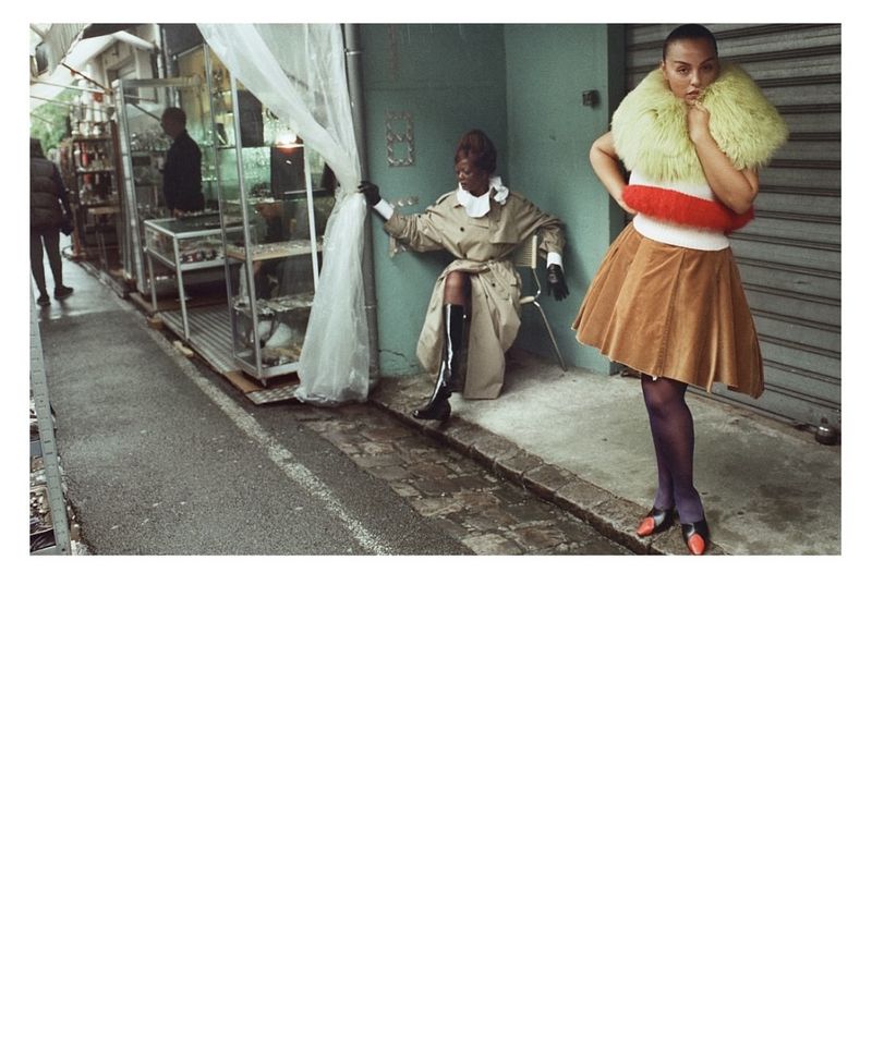Two fashion models pose outdoors, one sitting in a trench coat and the other standing in a yellow fur collar and brown skirt.