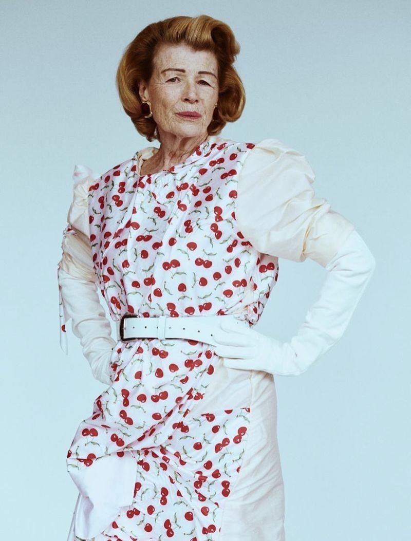 Elderly woman in a cherry-patterned dress with white belt and gloves poses in a studio with a confident expression, wearing gold earrings.