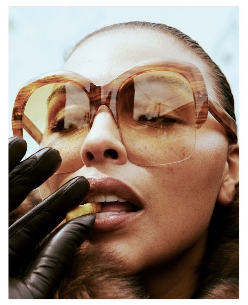 Close-up portrait of Paloma Elsesser wearing oversized tinted sunglasses and black leather gloves eating a snack.