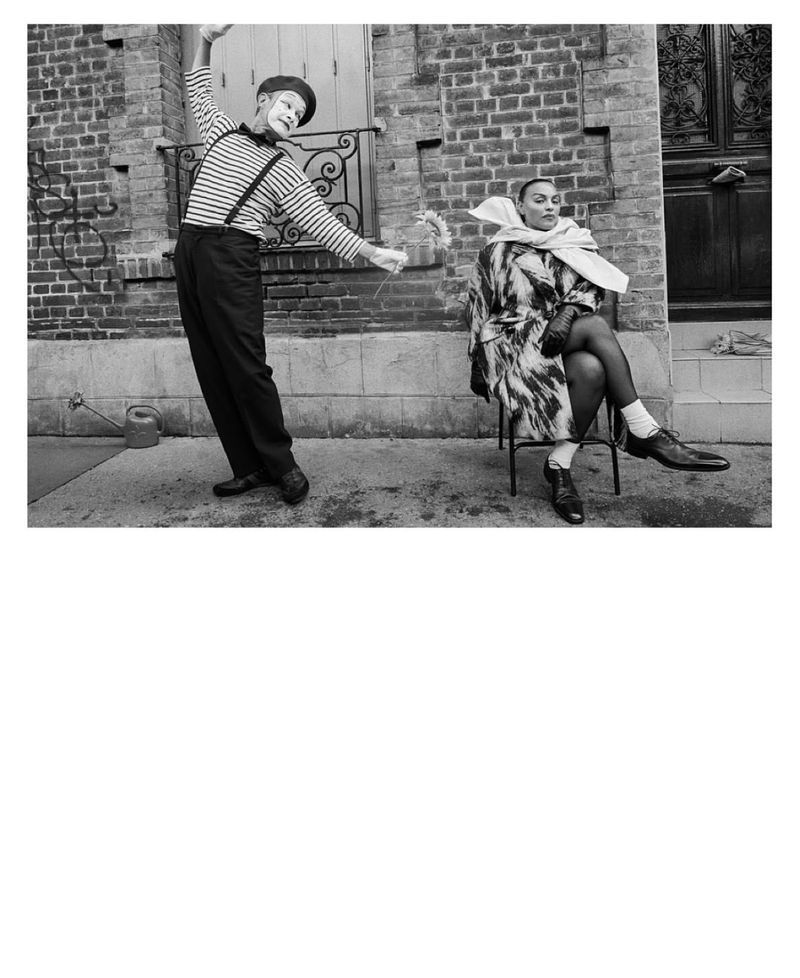 Monochrome portrait of a mime artist offering flowers to a woman seated on a chair outdoors in front of a brick wall.