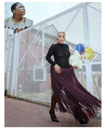 Paloma Elsesser in a black sheer top and dramatic purple fringe skirt holds a mannequin bust against a white mesh fence.