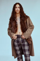 Model with long curly hair wearing a brown fur-trim coat and plaid trousers against a pale blue background