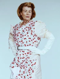 Mature woman posing with hands on hips wearing a cherry print dress and long white gloves against a light blue studio backdrop.