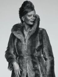 Black and white portrait of a woman with a high bouffant hairstyle wearing a thick belted fur coat against a plain studio background.