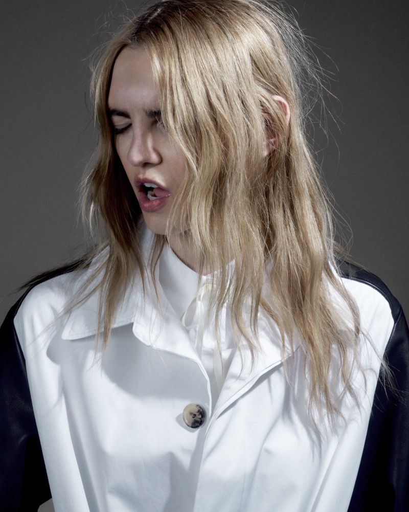 Portrait of a blonde adult woman in a white jacket with black sleeves, shot in a photo studio with a grey background.