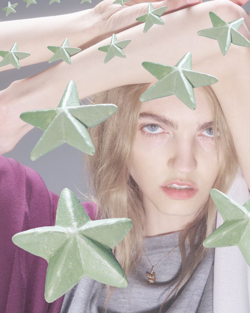 Young woman with blond hair poses with light green star objects; the scene features soft lighting and subdued, pastel color grading.
