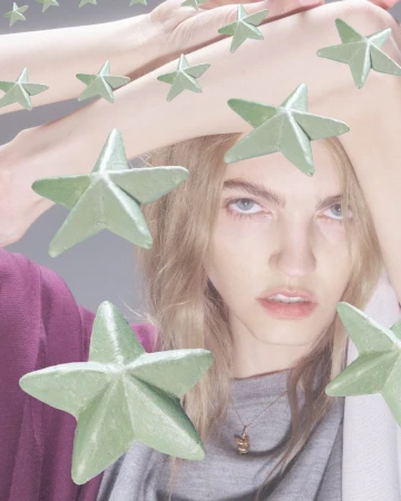 Young woman with blond hair poses with light green star objects; the scene features soft lighting and subdued, pastel color grading.