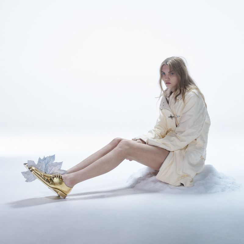 Fashion shot of a fair-skinned young woman seated on a cloud wearing a cream-colored jacket and shiny gold high-heeled shoes.
