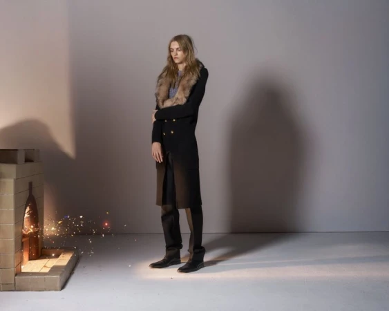 A tall woman in a dark coat with a fur collar poses indoors next to a modern-looking fireplace-like structure, facing right, against a neutral backdrop.