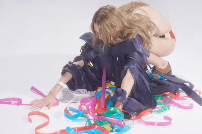 Two models pose in a studio with colorful streamers. One is wearing a blue and black costume, and the other is in red and white.