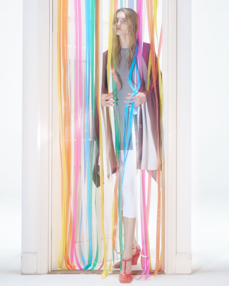 Full shot of a fair-skinned young adult woman in semi-sheer brown coat, t-shirt, white pants, and red T-strap heels behind colorful ribbons.
