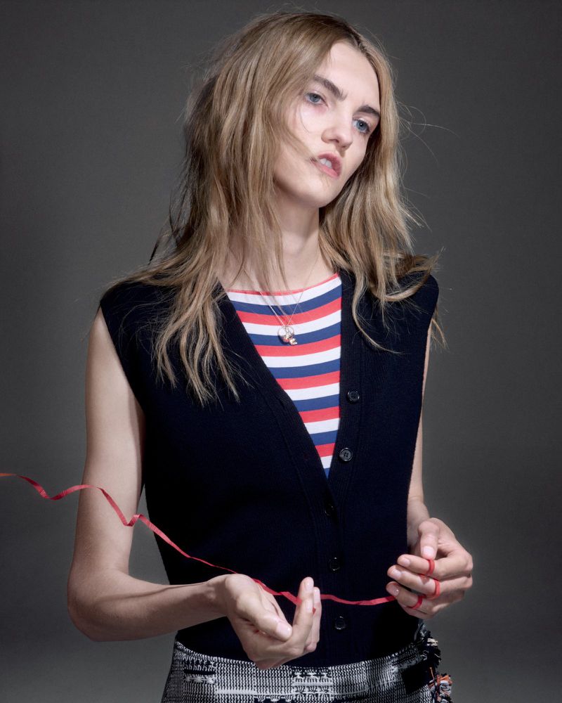 Stylish portrait of a young adult woman with blonde hair, wearing a striped top and vest, holding a red string indoors against a gray backdrop.
