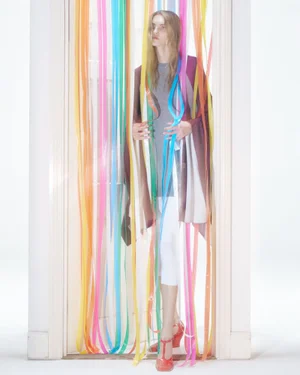 Model standing behind a doorway hung with colorful ribbon streamers in a white studio setting