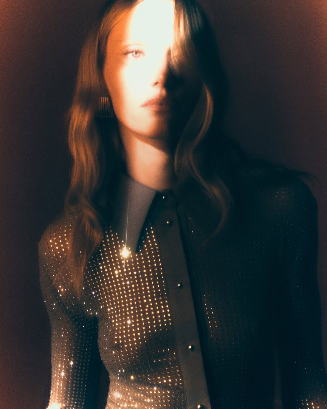 Close-up of a fair-skinned woman with wavy hair, wearing a sparkling, collared top in a studio setting with dramatic lighting.