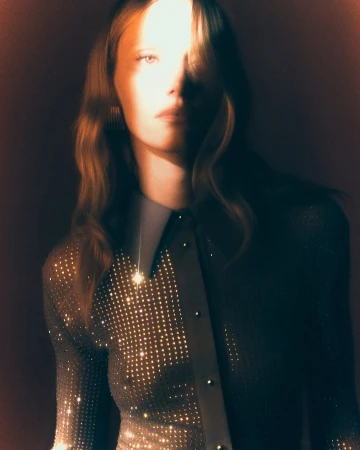 Close-up of a fair-skinned woman with wavy hair, wearing a sparkling, collared top in a studio setting with dramatic lighting.