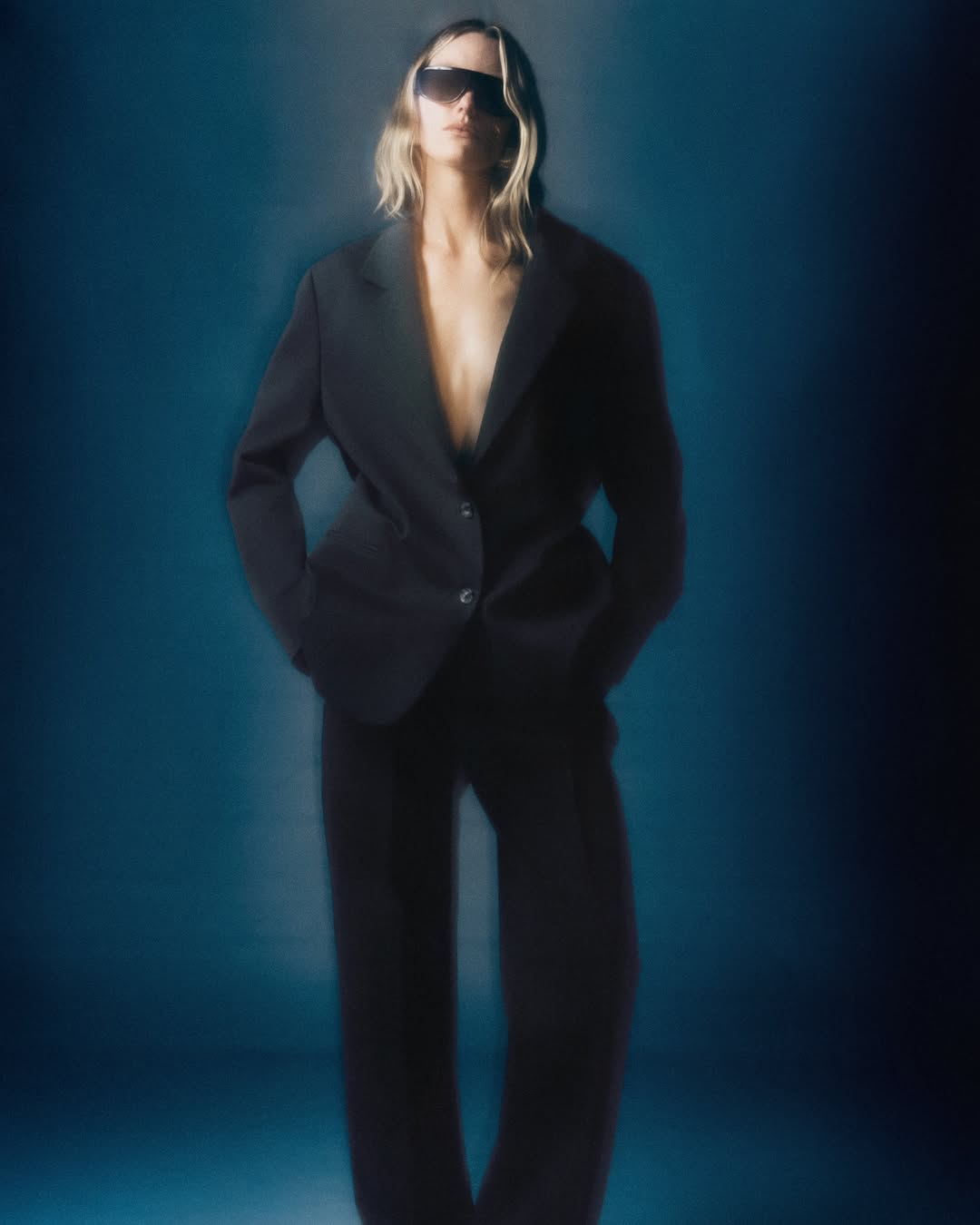 Fashion photo of a woman wearing a black pantsuit and sunglasses, standing against a gradient blue background in a studio setting.