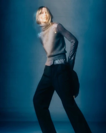Fashion shot of a woman in a grey turtleneck sweater with sequin sleeves and black pants against a dark teal-blue background.
