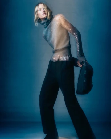 Model in translucent turtleneck sweater and black trousers holding a dark handbag against a moody blue gradient background.