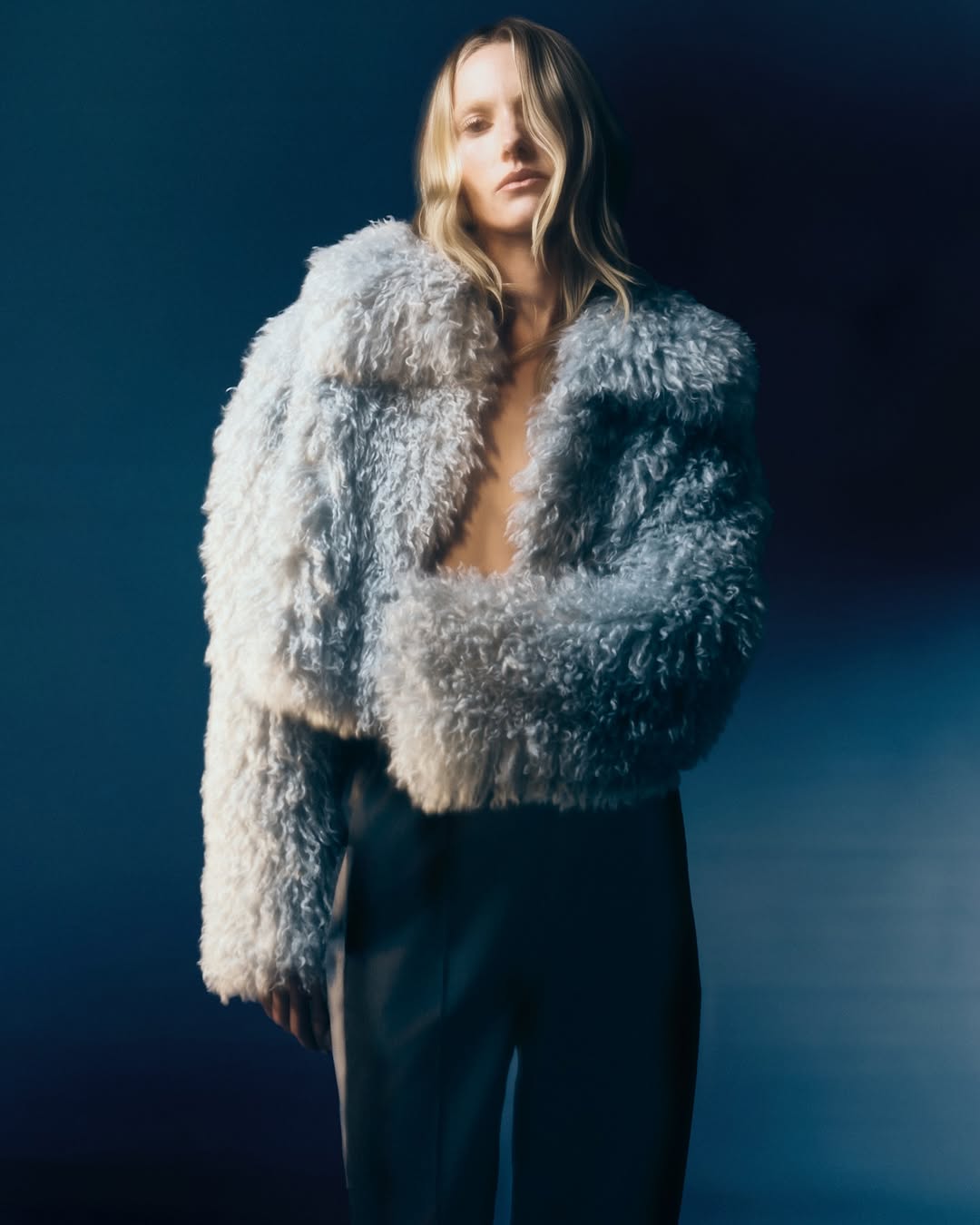A blonde woman poses in a fuzzy blue jacket and pants against a gradient blue background in a fashion studio shot.