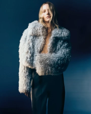 A blonde woman poses in a fuzzy blue jacket and pants against a gradient blue background in a fashion studio shot.