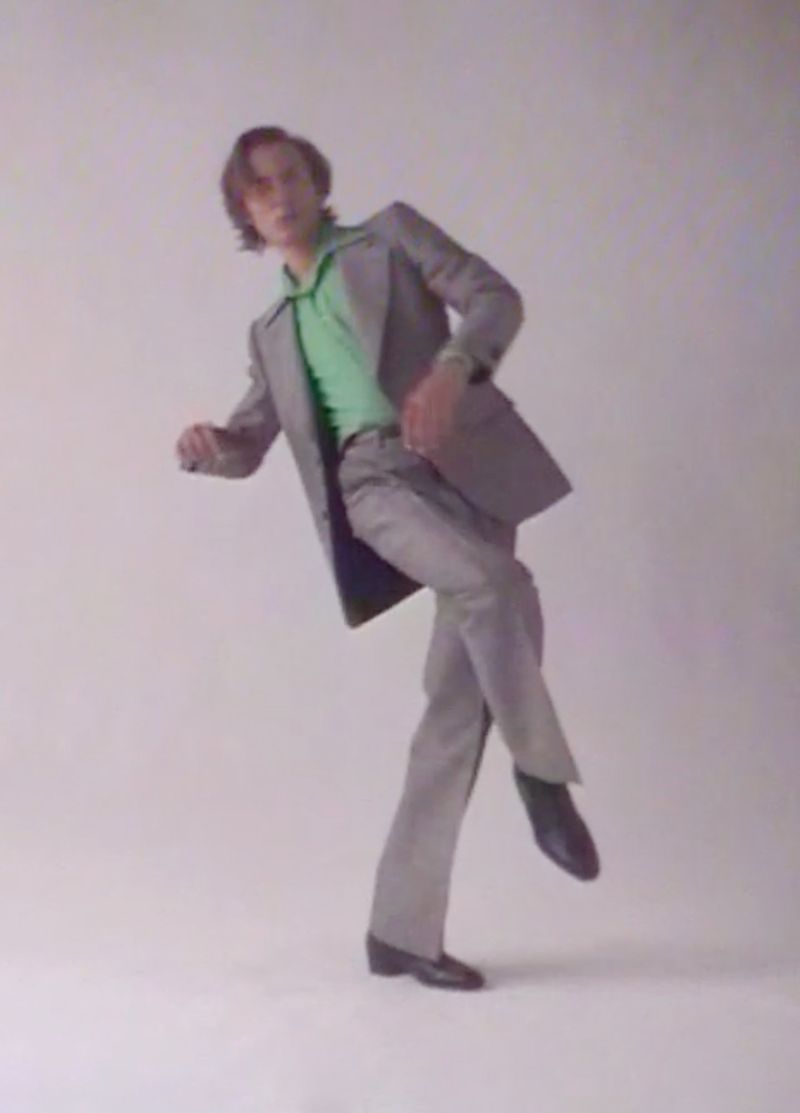 Adult male in a light grey suit and green shirt poses with one leg raised in a photo studio against a plain background.