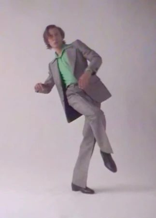 Adult male in a light grey suit and green shirt poses with one leg raised in a photo studio against a plain background.