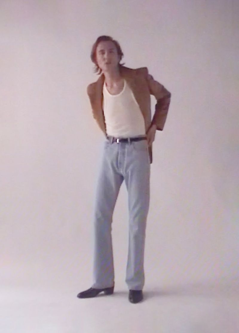 Full-length studio portrait of a young adult man in a tan blazer, cream tank top, blue jeans, and black shoes against a white backdrop.