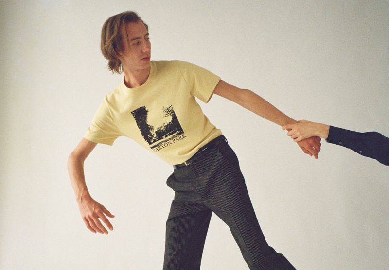 A slender man in a graphic tee holds hands with another person in a minimalist studio, emphasizing fashion and interaction.