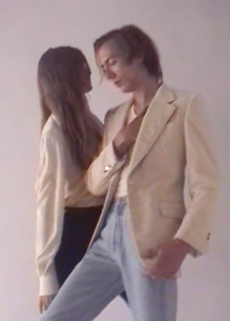 Couple poses together in a photo studio with a man in a cream suit jacket and light jeans in the foreground, and a woman in background.