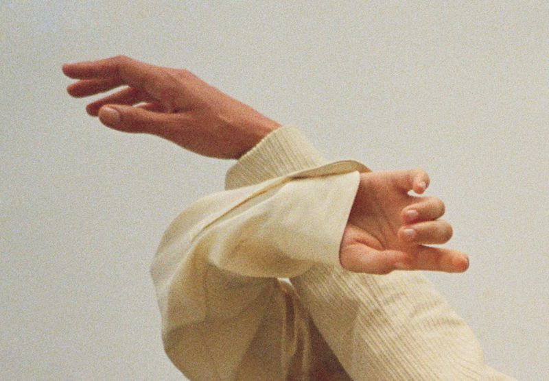 Close-up view of two hands and arms, one extended upward, one angled down, wearing a textured, neutral-colored garment against a pale background.
