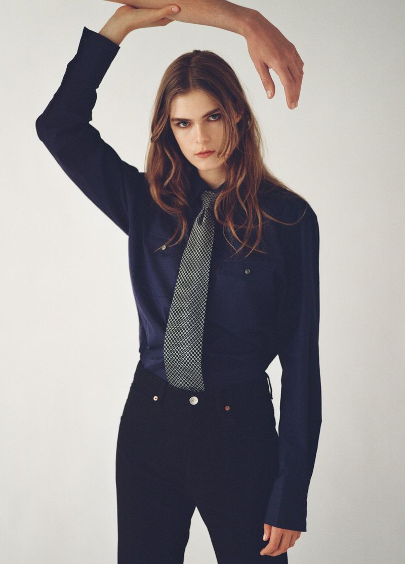 A woman poses indoors wearing a navy shirt and black jeans, accessorized with a patterned tie and an outstretched hand above her.