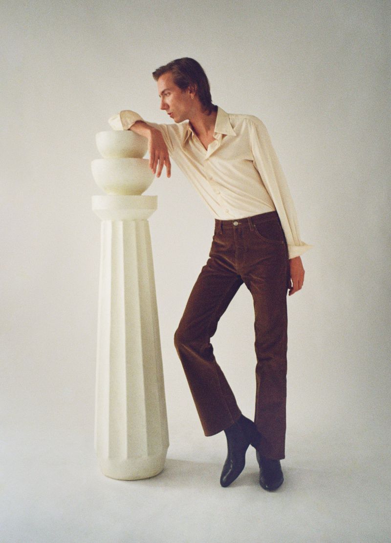 A slender, light-skinned man in 70's attire leans against a fluted column topped with ceramic bowls in a soft, diffused indoor lighting.