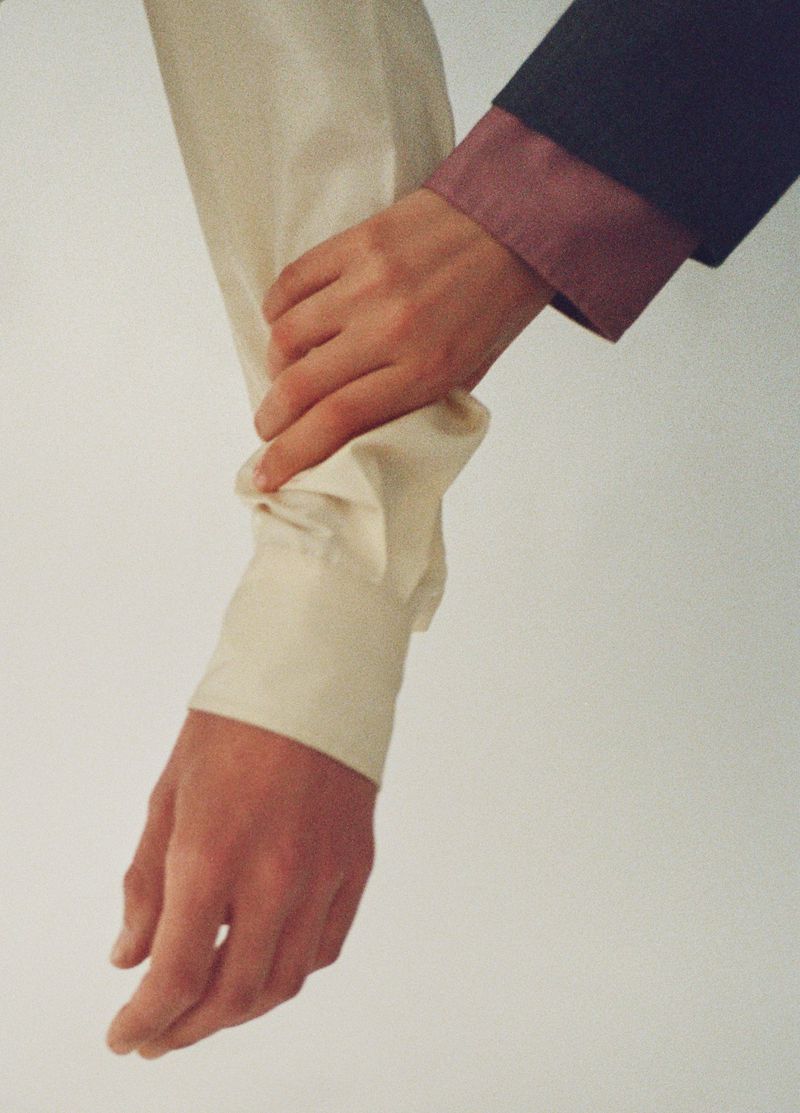 Close-up of two hands, one adjusting the cuff of a dress shirt, showcasing details in fabric and fit against a white backdrop.