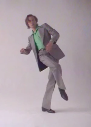 Model Paul Hameline posing dynamically on one leg in a grey tailored suit and green shirt against a white studio background.