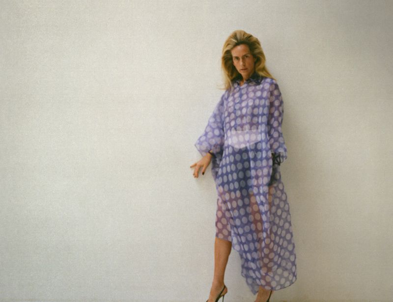 Woman in a sheer, polka dot, lilac dress poses against a white wall, showcasing a blend of fashion and studio photography.