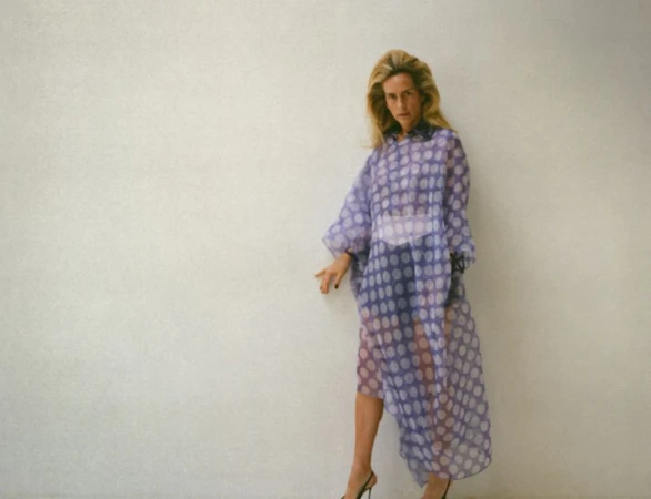 Woman in a sheer, polka dot, lilac dress poses against a white wall, showcasing a blend of fashion and studio photography.