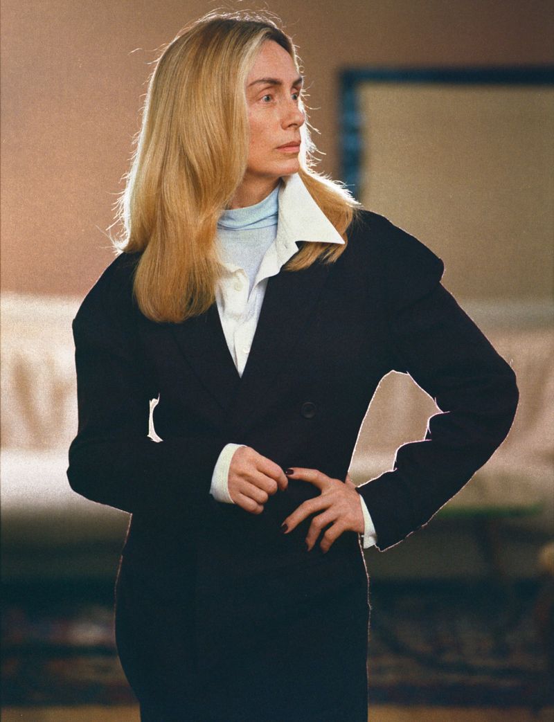 Portrait of a blonde woman in a black jacket over a light blue turtleneck and white shirt, captured in a medium shot indoors.