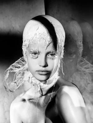 Black and white editorial portrait of a woman wearing a textured white headpiece and white feathery eyelashes with shadow play.