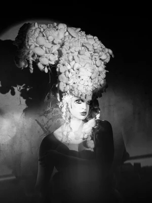 Black and white portrait of a woman wearing an elaborate towering mushroom-like sculptural headpiece with dramatic spotlighting.